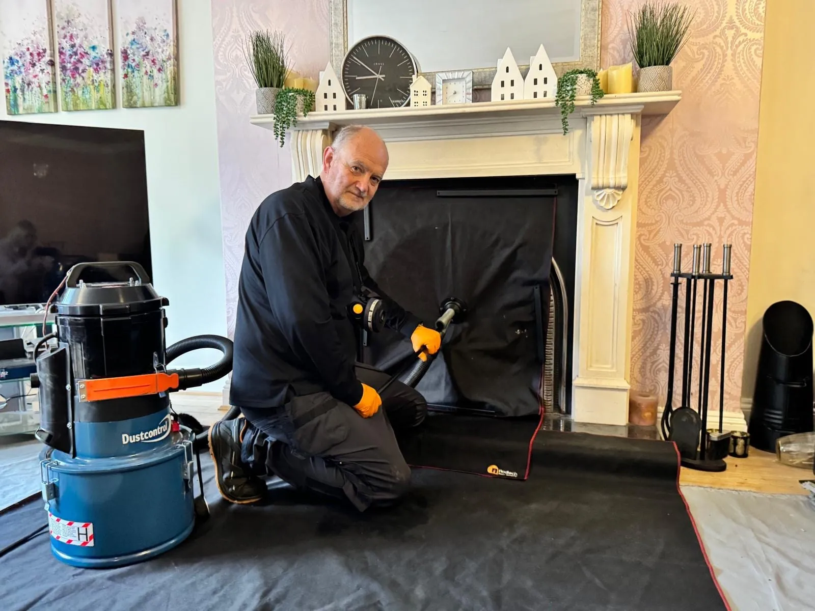 Chimney sweep kneeling at a fireplace sealed with a sheet, with a vacuum and dust sheets protecting the room